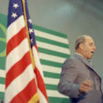 Gaylord Nelson speaking at a podium during Earth Week with an American flag in the foreground, and the Earth Day flag behind it.