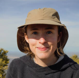 Close-up portrait of a young woman wearing a large khaki sun hat and a gray hoodie against a background of blue sky and green desert shrubs.